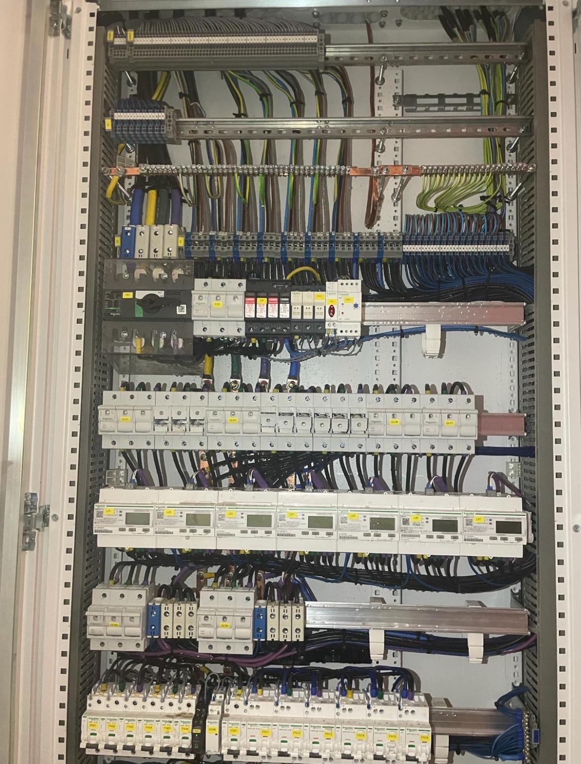 Rozdzielnica elektryczna w szafie sterowniczej z precyzyjnie ułożonymi przewodami, zabezpieczeniami i licznikami energii elektrycznej. Widok od frontu.