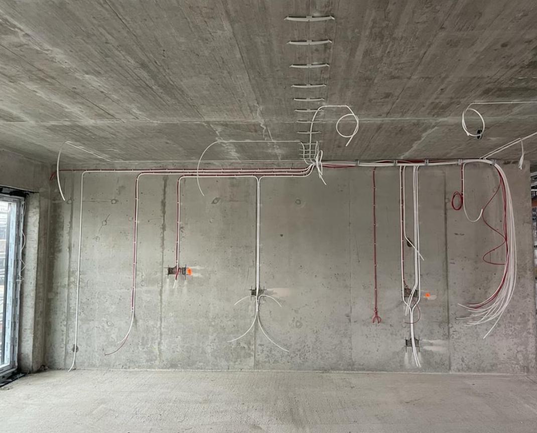 Surowy stan: Instalacja elektryczna w nowym budynku z betonowymi ścianami i widocznymi przewodami w kolorze białym i czerwonym. Przewody poprowadzone po suficie i ścianach.