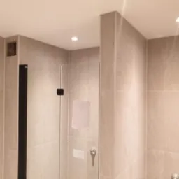 Beżowa łazienka z kabiną prysznicową i czarnymi detalami. Widoczna wnęka w ścianie, jasne płytki i oświetlenie punktowe.