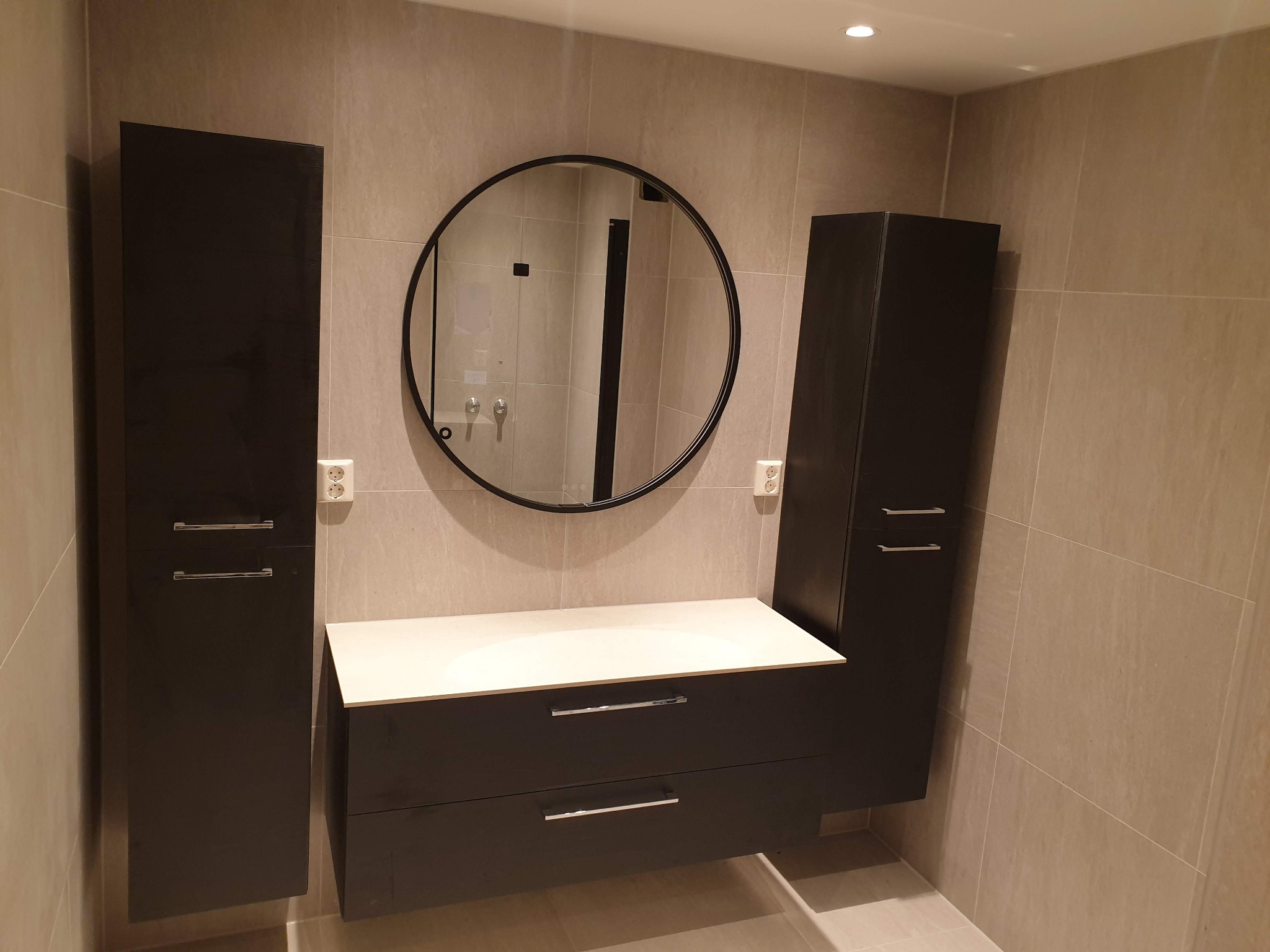 Nowoczesna łazienka z szarymi płytkami, okrągłym lustrem w czarnej ramie, szafką pod umywalką i wysokimi szafkami po bokach. Minimalistyczny design.