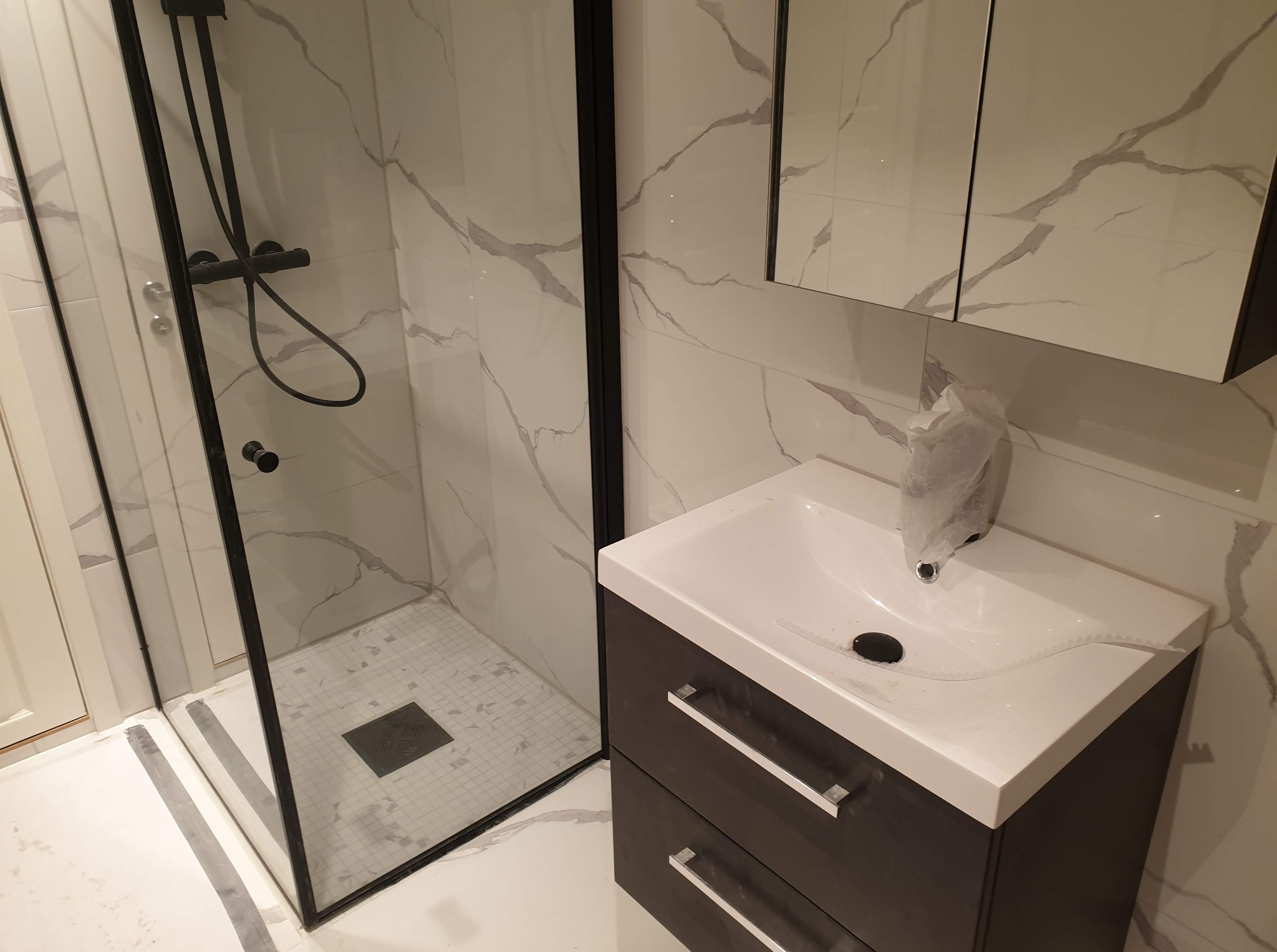 Nowoczesna łazienka z kabiną prysznicową typu walk-in, umywalką z szafką w kolorze grafitu i białymi płytkami imitującymi marmur z szarymi żyłami.