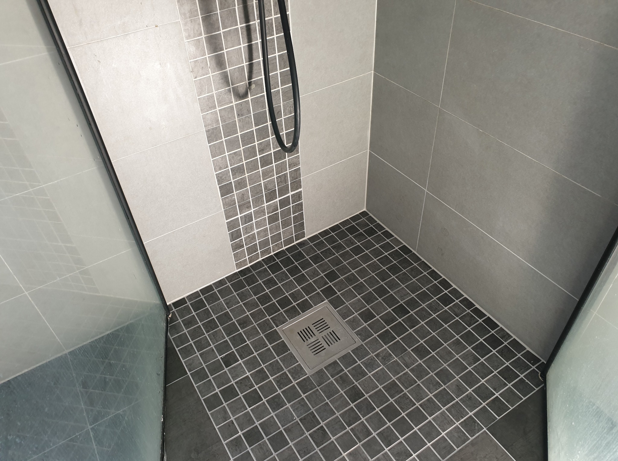 Wykończona kabina prysznicowa z szarymi płytkami, mozaiką i odpływem liniowym. Widoczne szklane drzwi kabiny i czarny wąż prysznicowy. Ukończony remont łazienki.