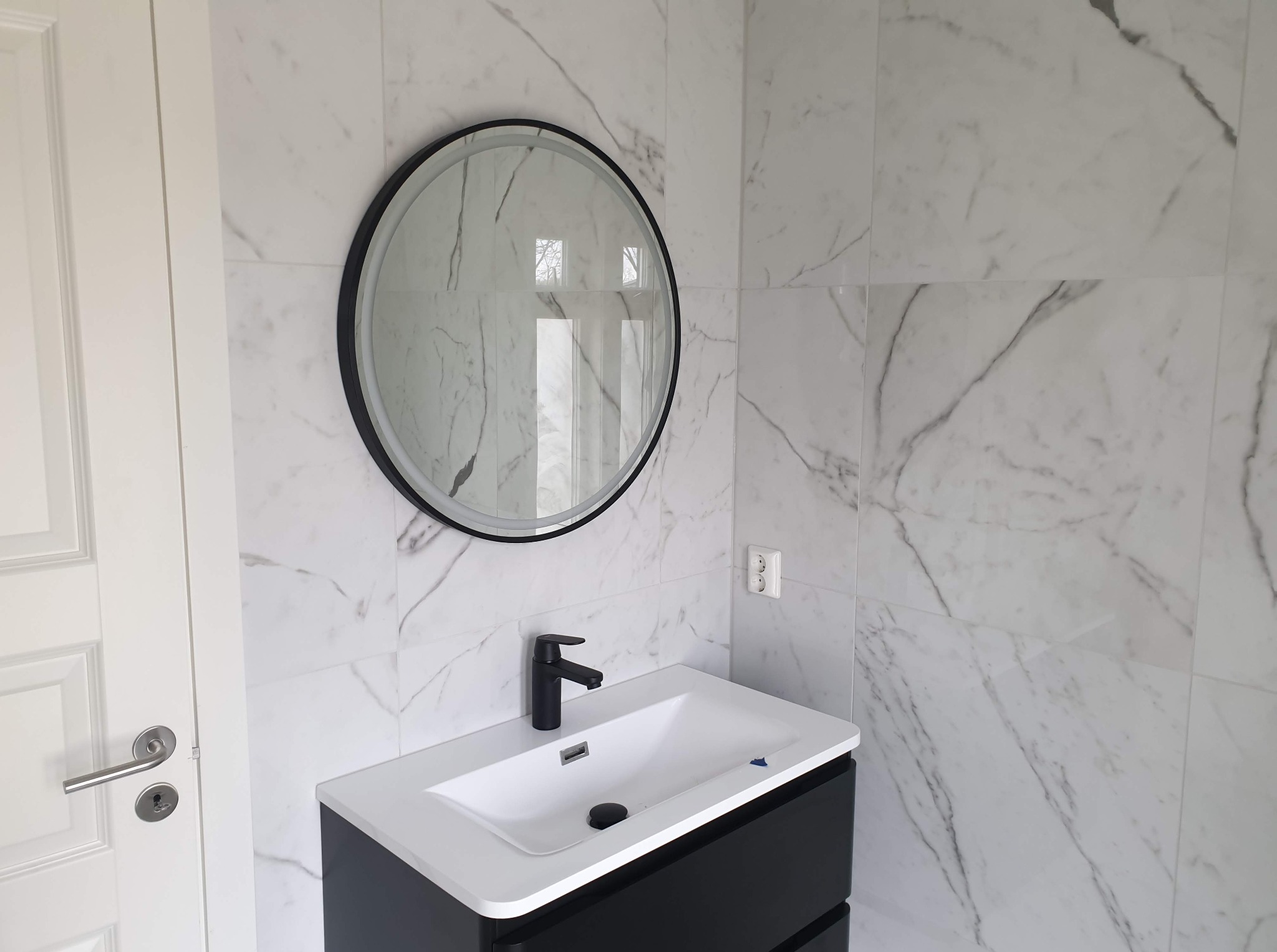 Nowoczesna łazienka z umywalką na szafce w kolorze czarnym, okrągłym lustrem w czarnej ramie i białymi płytkami imitującymi marmur na ścianie.