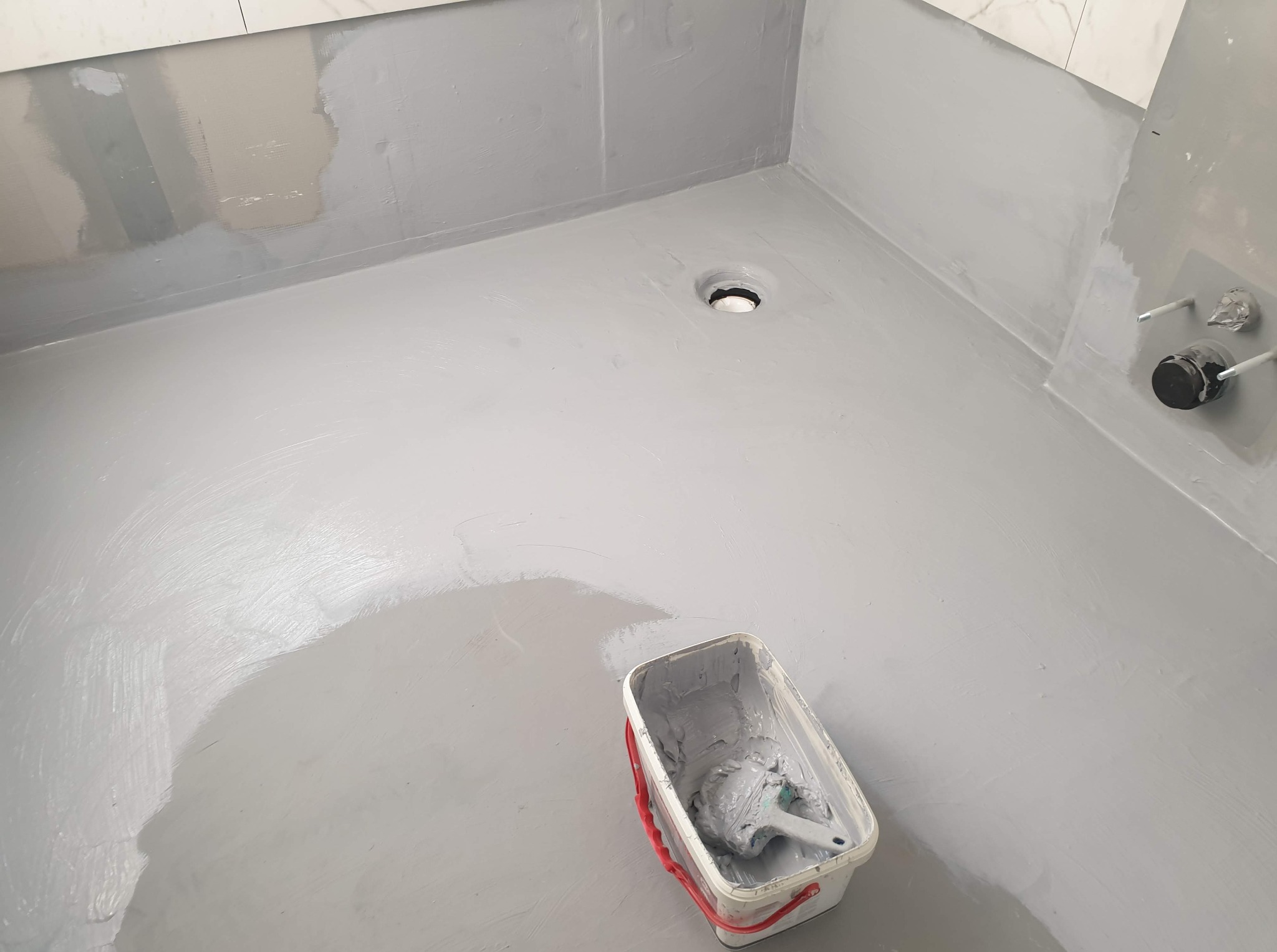 Hydroizolacja podłogi w łazience, szara masa uszczelniająca, wiadro z pędzlem w rogu, widoczne fragmenty płytek na ścianie i odpływ liniowy.