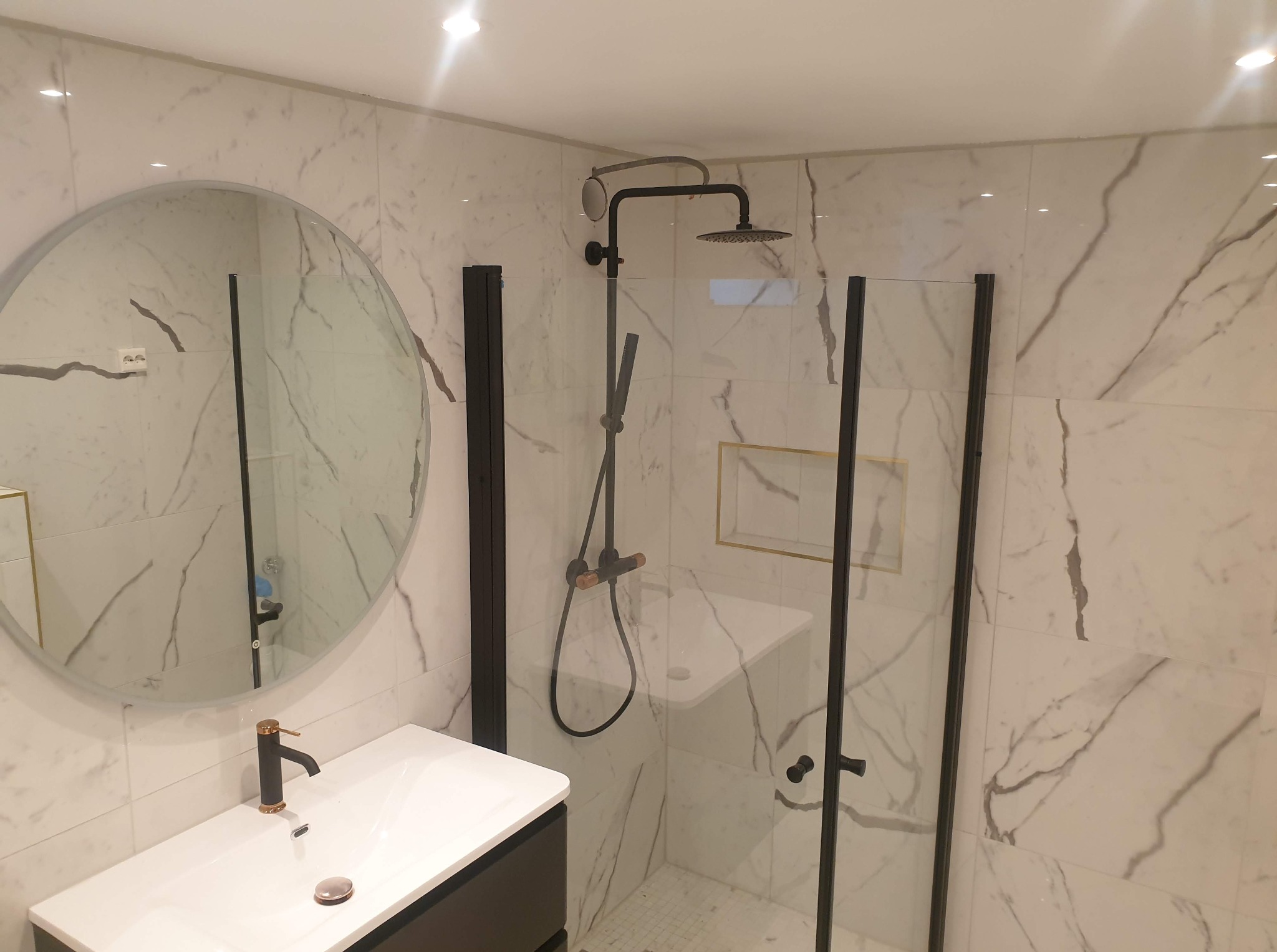 Nowoczesna łazienka z kabiną prysznicową typu walk-in, marmurowymi płytkami i okrągłym lustrem. Czarna armatura i biała umywalka z szafką podwieszaną.