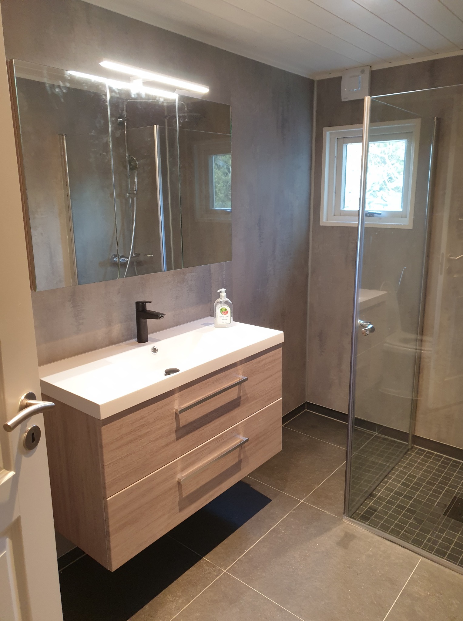 Nowoczesna łazienka z drewnianą szafką pod umywalką i kabiną prysznicową. Szare płytki na ścianach i podłodze, minimalistyczny design.