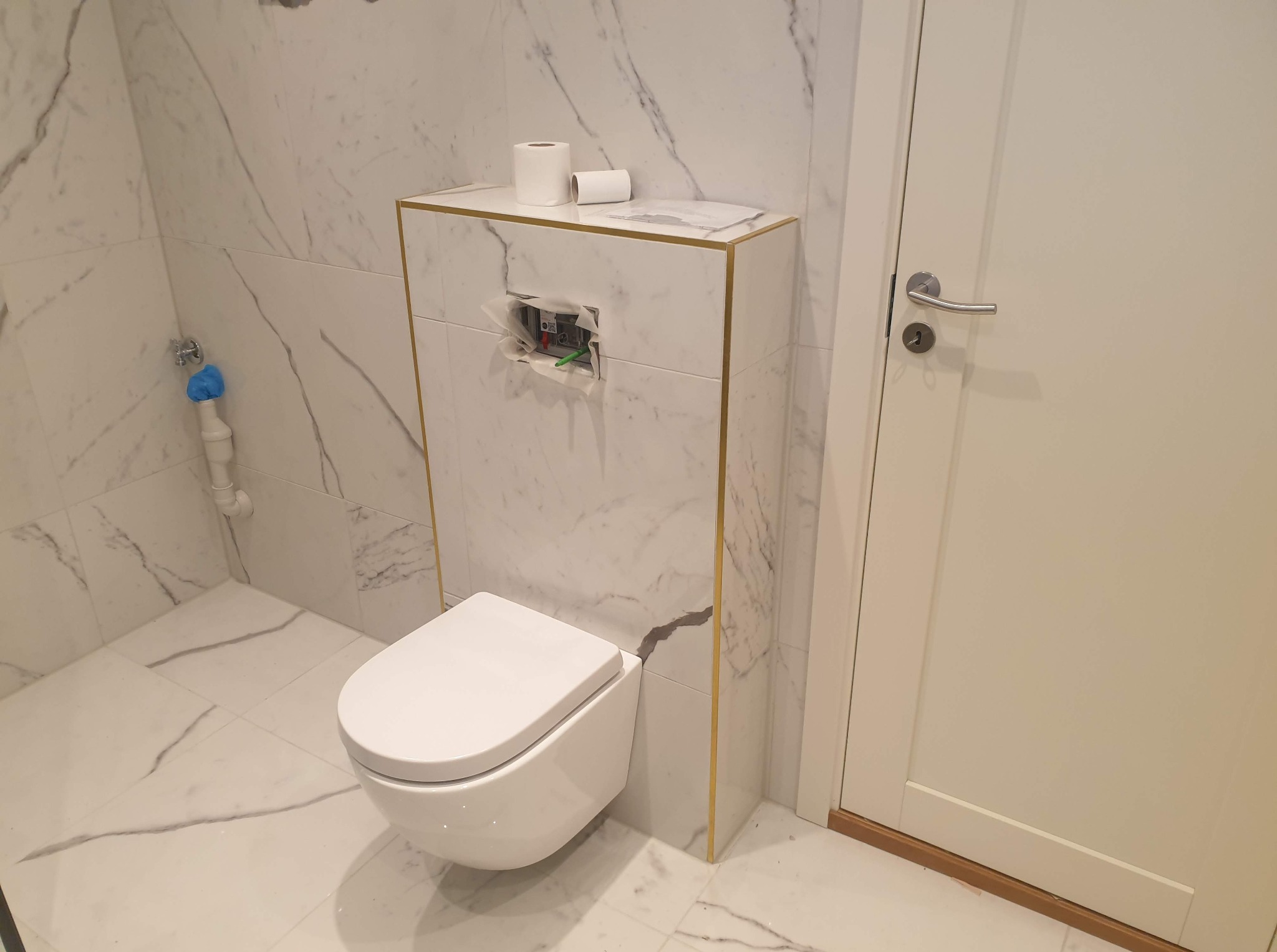 Nowoczesna toaleta podwieszana w łazience wykończonej płytkami imitującymi marmur, z widoczną instalacją i rolkami papieru toaletowego na obudowie stelaża.