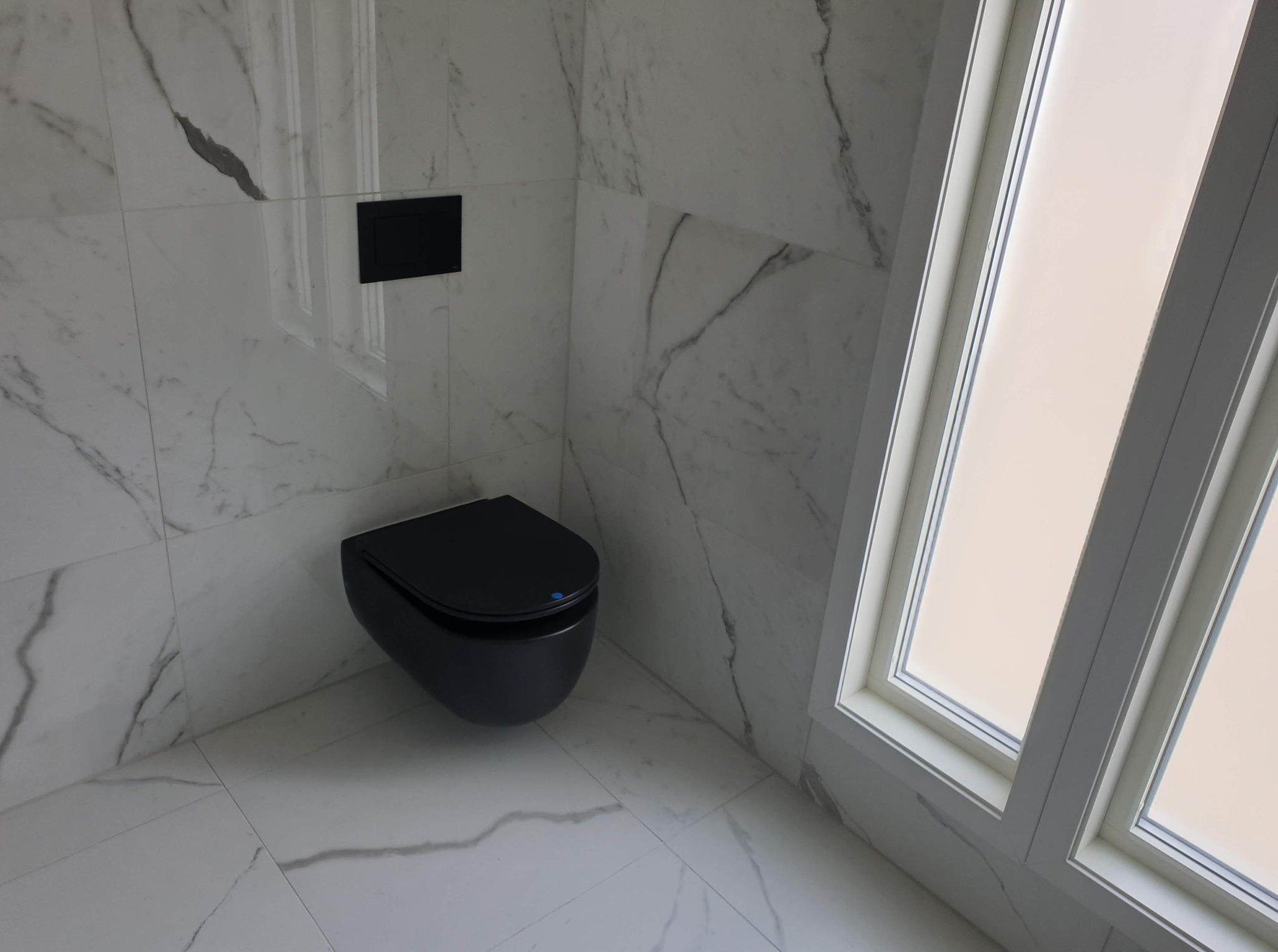 Elegancka łazienka: czarna, podwieszana toaleta na tle marmurowych płytek i okna z białymi ramami. Minimalistyczny design z dbałością o detale.
