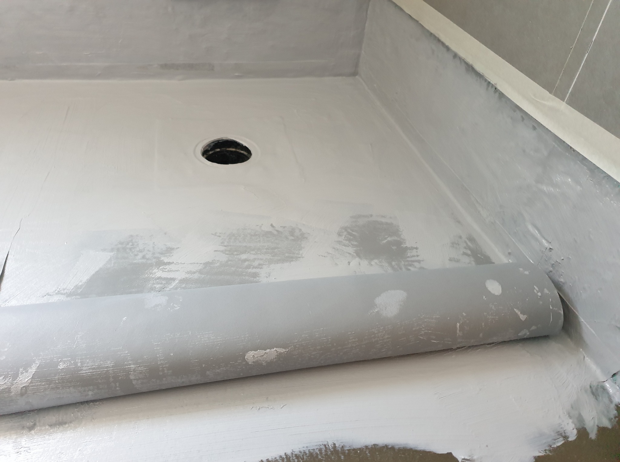 Hydroizolacja brodzika w łazience z użyciem folii w płynie i taśmy uszczelniającej. Widoczny odpływ liniowy i zabezpieczone narożniki. Szare płytki w tle.