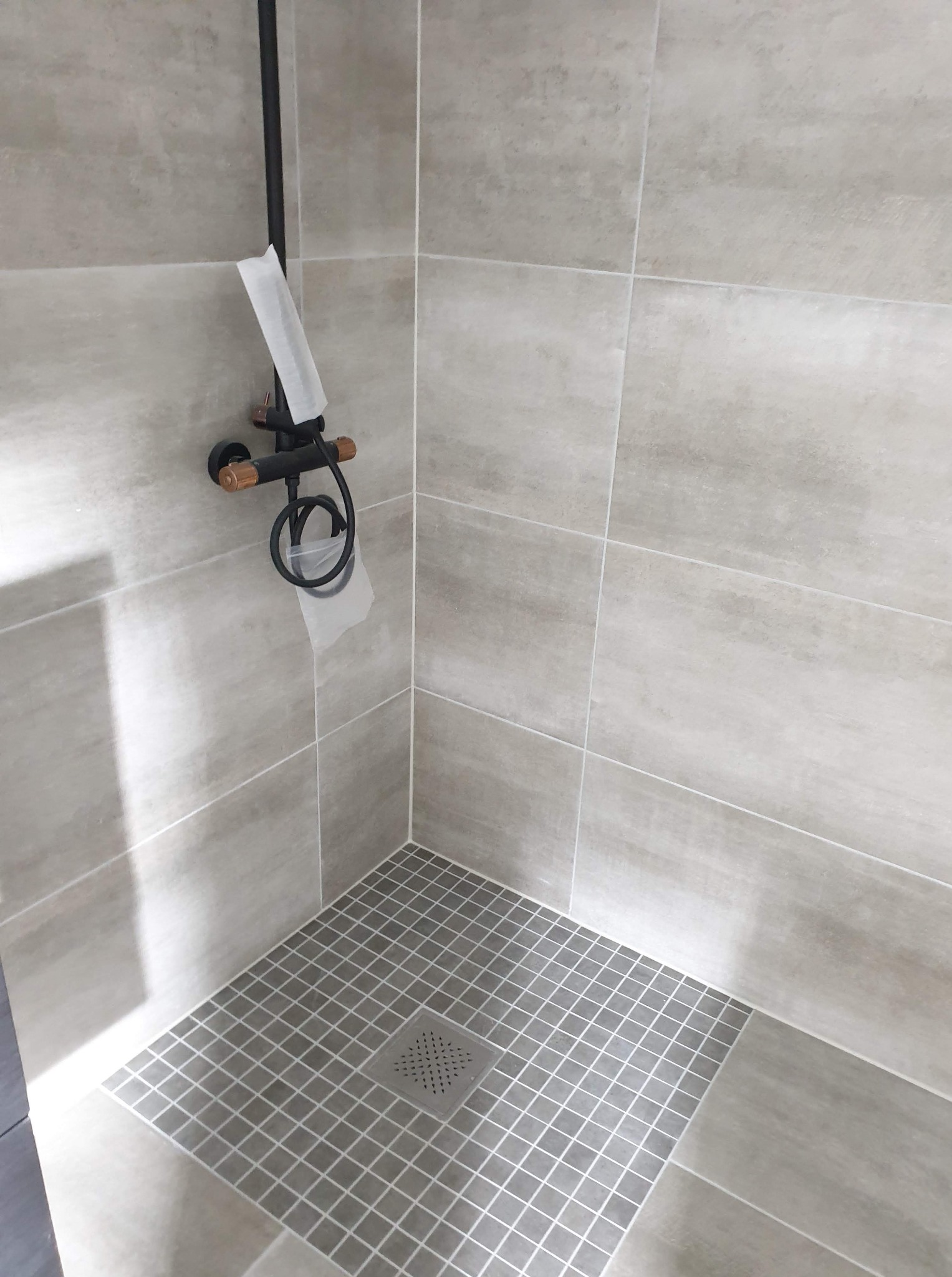 Wykończona kabina prysznicowa z szarymi płytkami, czarną armaturą z elementami drewna i odpływem liniowym. Minimalistyczny design, neutralna kolorystyka.