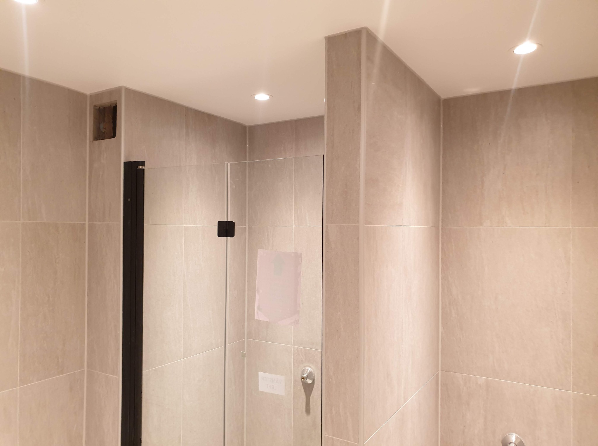 Beżowa łazienka z kabiną prysznicową i czarnymi detalami. Widoczna wnęka w ścianie, jasne płytki i oświetlenie punktowe.