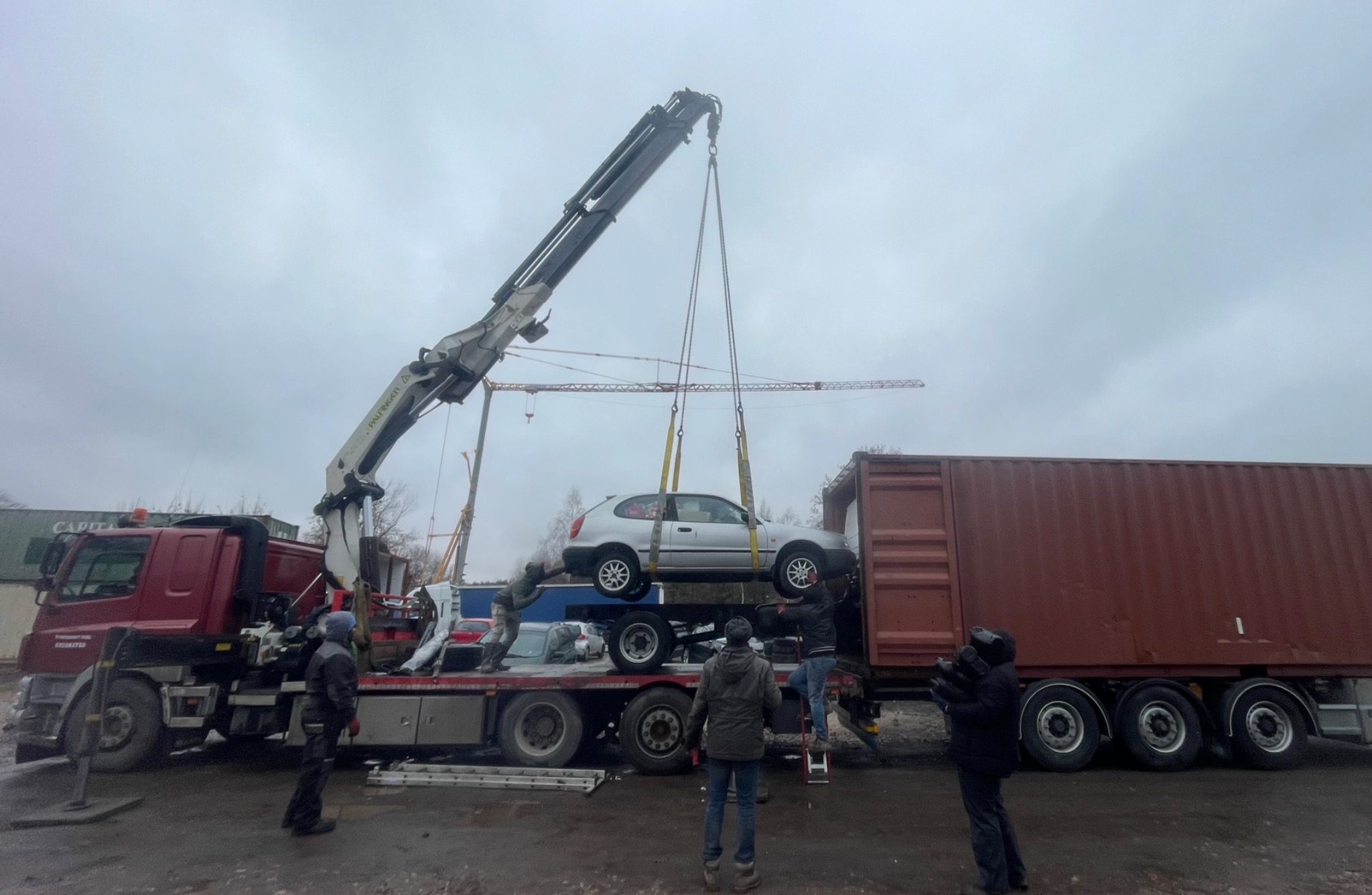 Samochód osobowy podnoszony dźwigiem w celu załadunku do kontenera transportowego na lawecie. Praca w toku, widoczni pracownicy i dźwig. Pochmurny dzień.