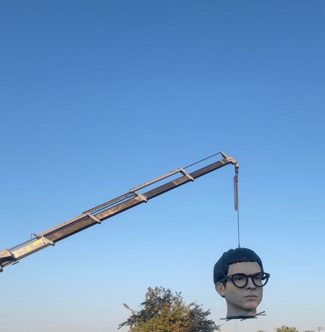 Dźwig podnosi dużą głowę manekina na tle błękitnego nieba. Nietypowe ujęcie z dołu. Widoczny fragment drzewa.