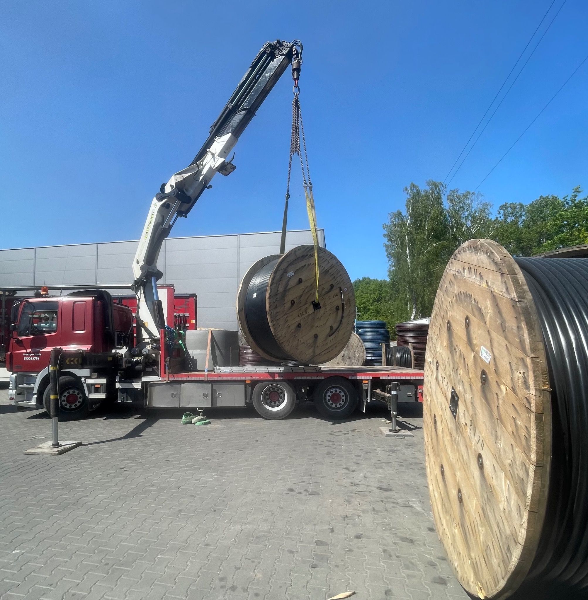 Żuraw samochodowy podnosi bęben z kablem na tle błękitnego nieba i zieleni. Widoczny czerwony samochód ciężarowy z żurawiem i fragmenty innych bębnów.