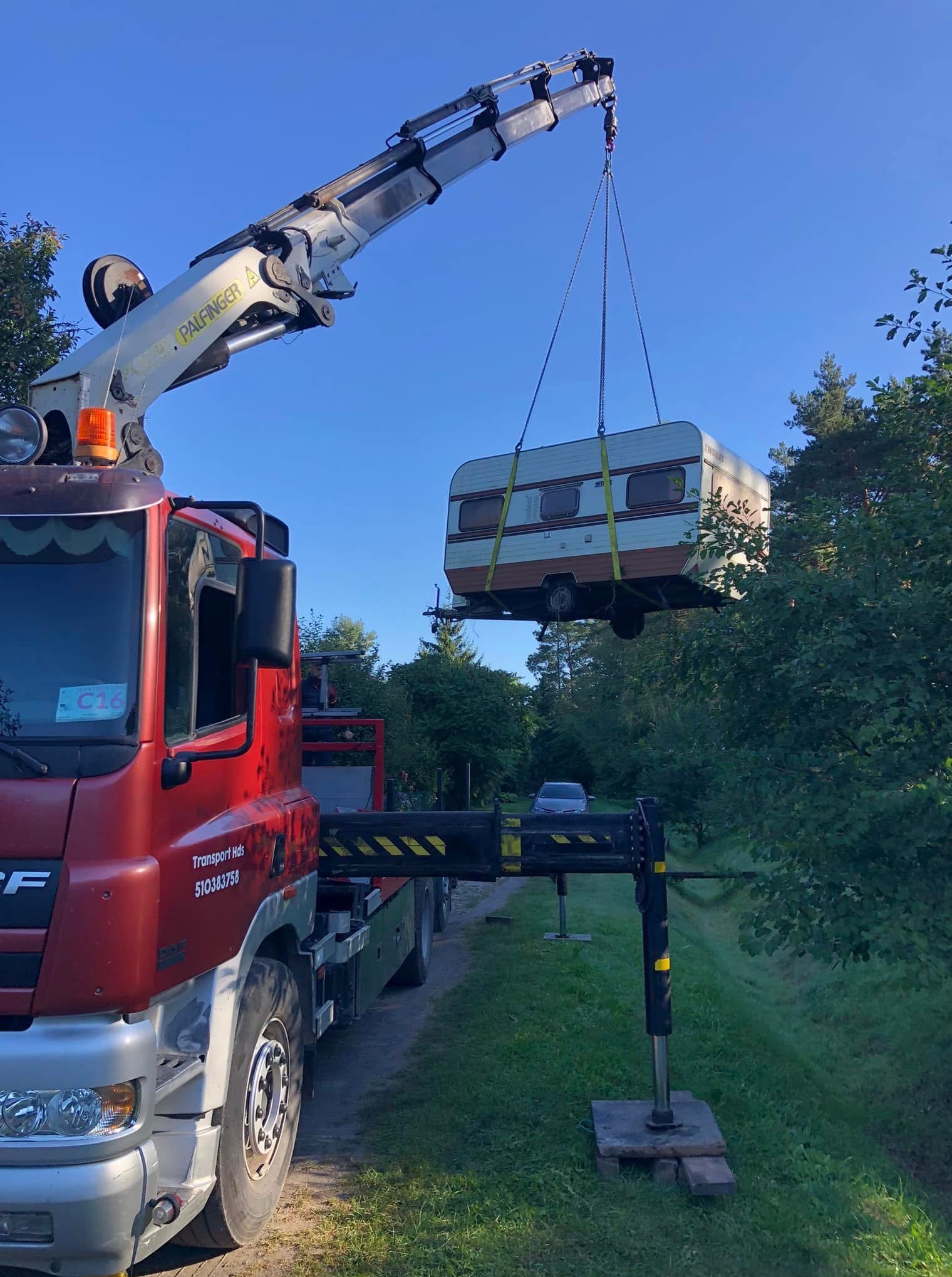 Czerwony dźwig Palfinger unosi przyczepę kempingową w zielonym otoczeniu. Podpory dźwigu na betonowych płytach. Widoczny numer telefonu na kabinie.