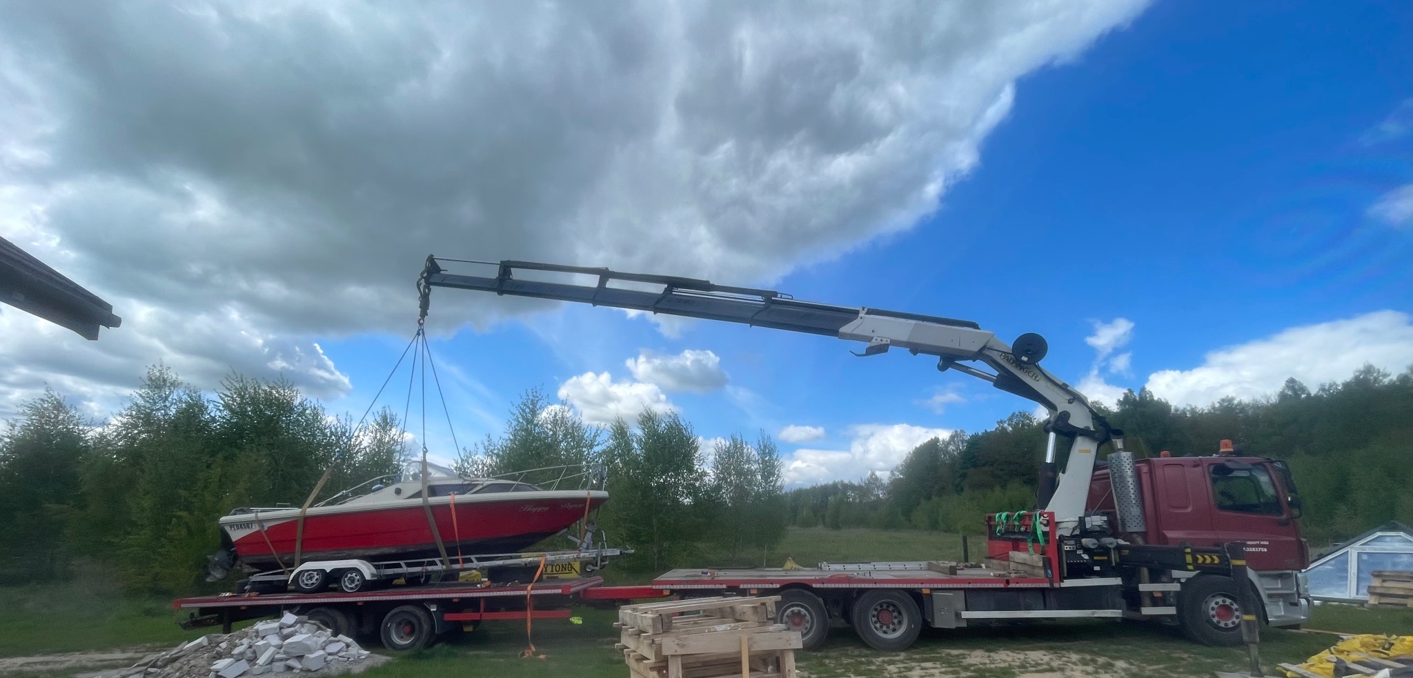 Czerwona łódź motorowa podnoszona dźwigiem samochodowym na tle zieleni i błękitnego nieba z chmurami. Widoczny fragment kabiny i przyczepy pod łodzią.