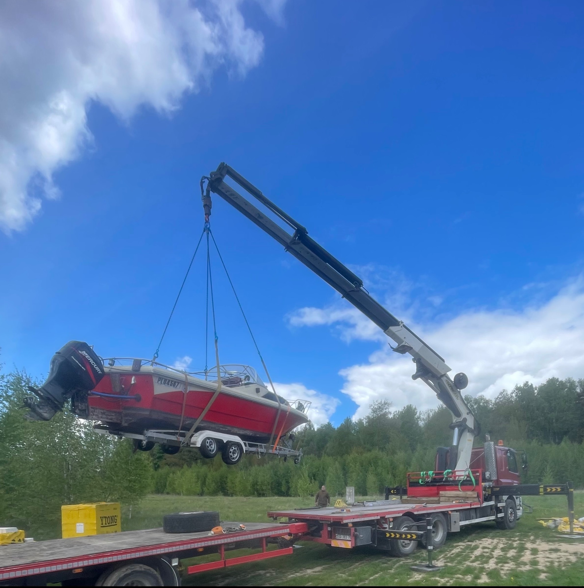 Czerwona łódź motorowa na przyczepie, podnoszona przez dźwig samochodowy z wysięgnikiem teleskopowym na tle błękitnego nieba z chmurami i zieleni drzew.
