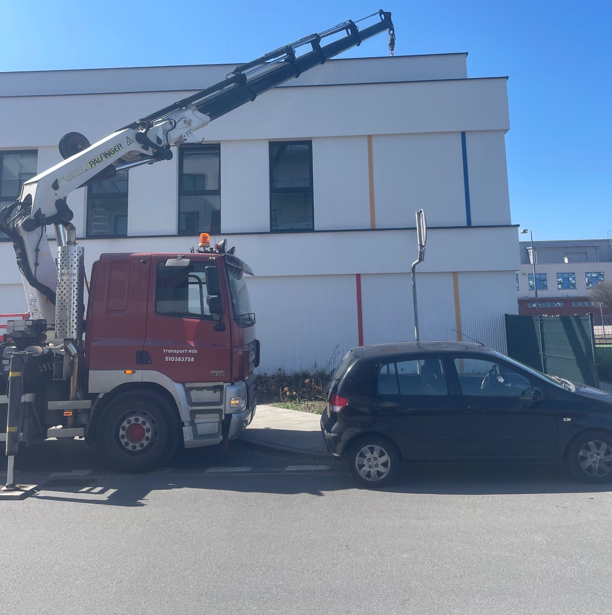 Ciężarówka z dźwigiem Palfinger zaparkowana obok czarnego samochodu osobowego na tle budynku z kolorowymi pasami. Widok z boku, jasne oświetlenie.