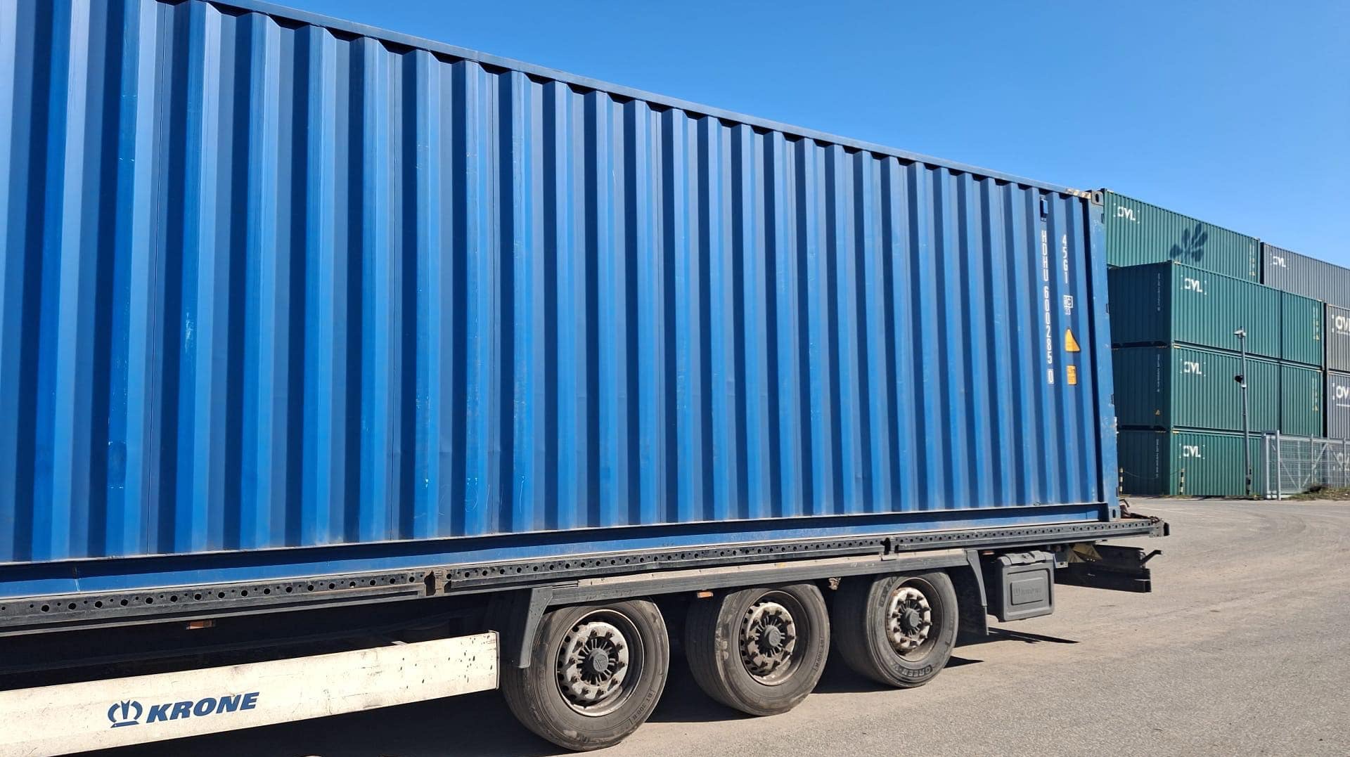 Niebieski kontener na naczepie ciężarówki, widok z boku na tle innych kontenerów i błękitnego nieba. Widoczne koła i fragment podwozia z logo Krone.
