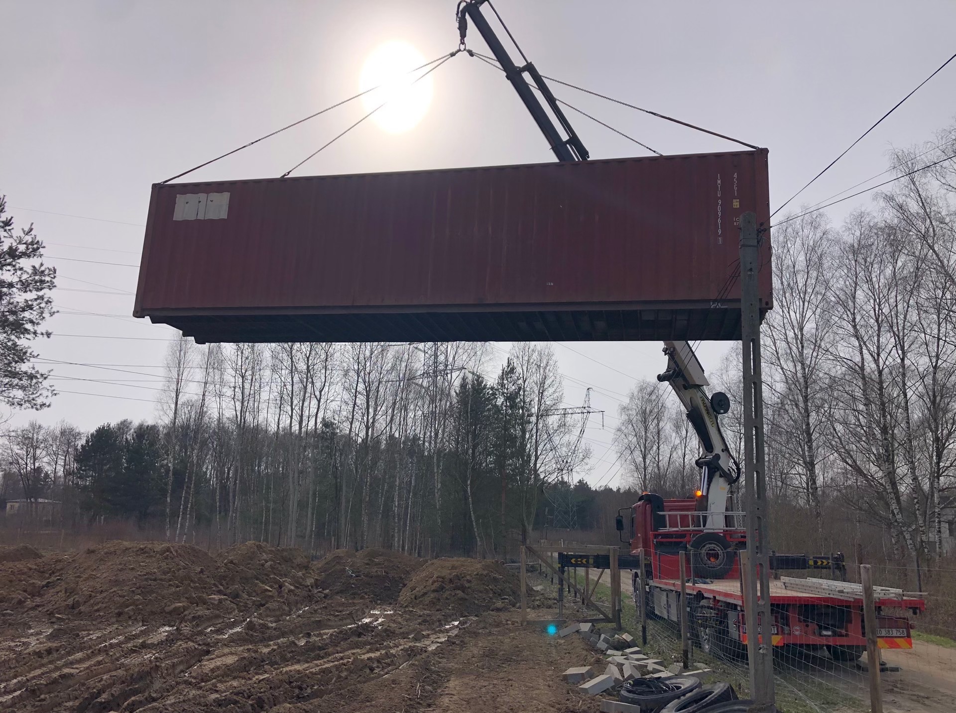 Dźwig samochodowy podnosi kontener w pobliżu terenu budowy. Widoczne drzewa i linia energetyczna w tle, pochmurne niebo.