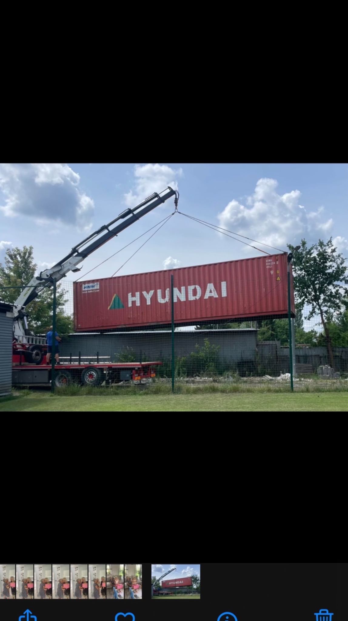 Czerwony kontener Hyundai podnoszony dźwigiem na tle zieleni i błękitnego nieba z chmurami. Widoczny fragment ciężarówki i operator dźwigu.