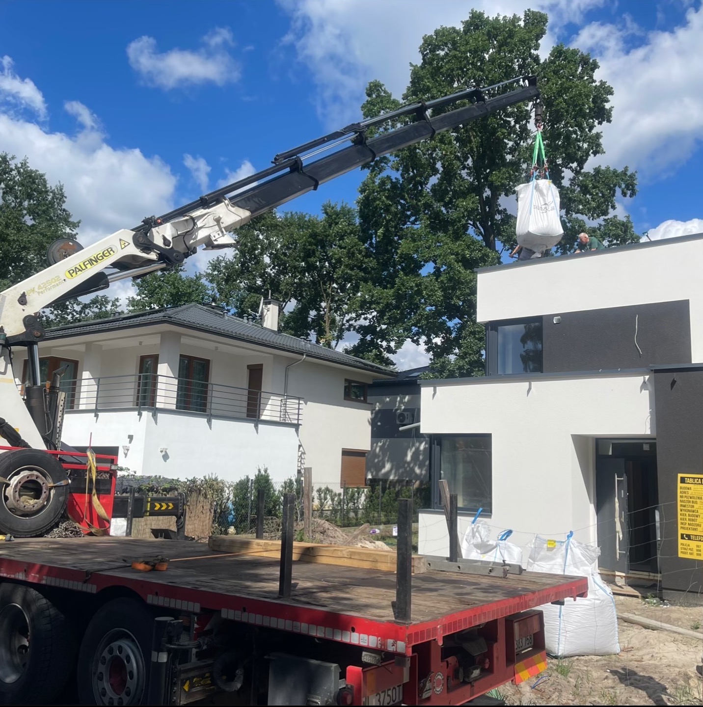 Dźwig Palfinger podnosi duży worek na dach nowoczesnego domu jednorodzinnego. Widoczna platforma transportowa, zieleń i błękitne niebo w tle.