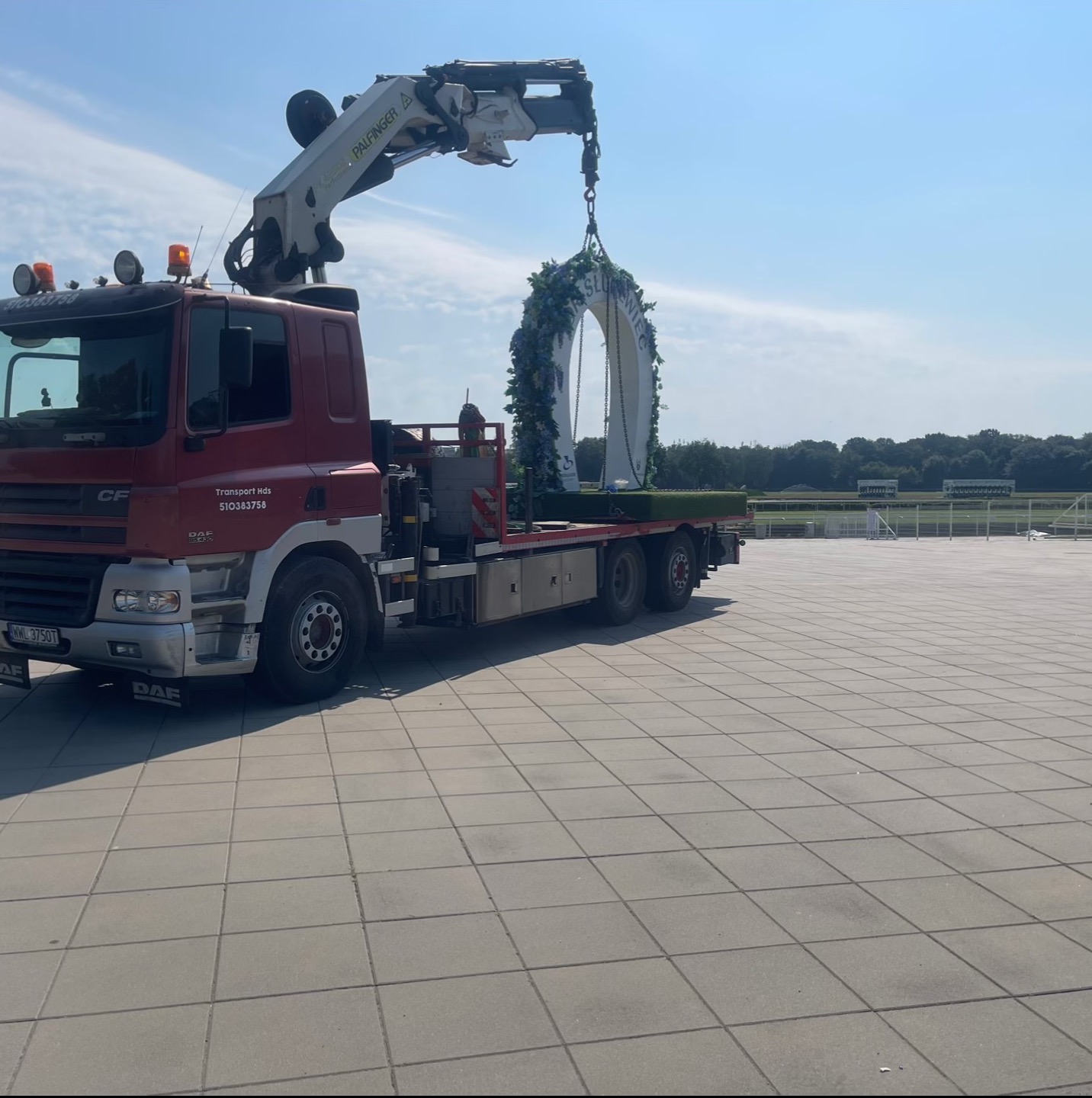 Czerwony dźwig samochodowy Palfinger transportuje ozdobny, biały łuk z kwiatami na tle błękitnego nieba i betonowego placu. Widoczny numer rejestracyjny pojazdu.