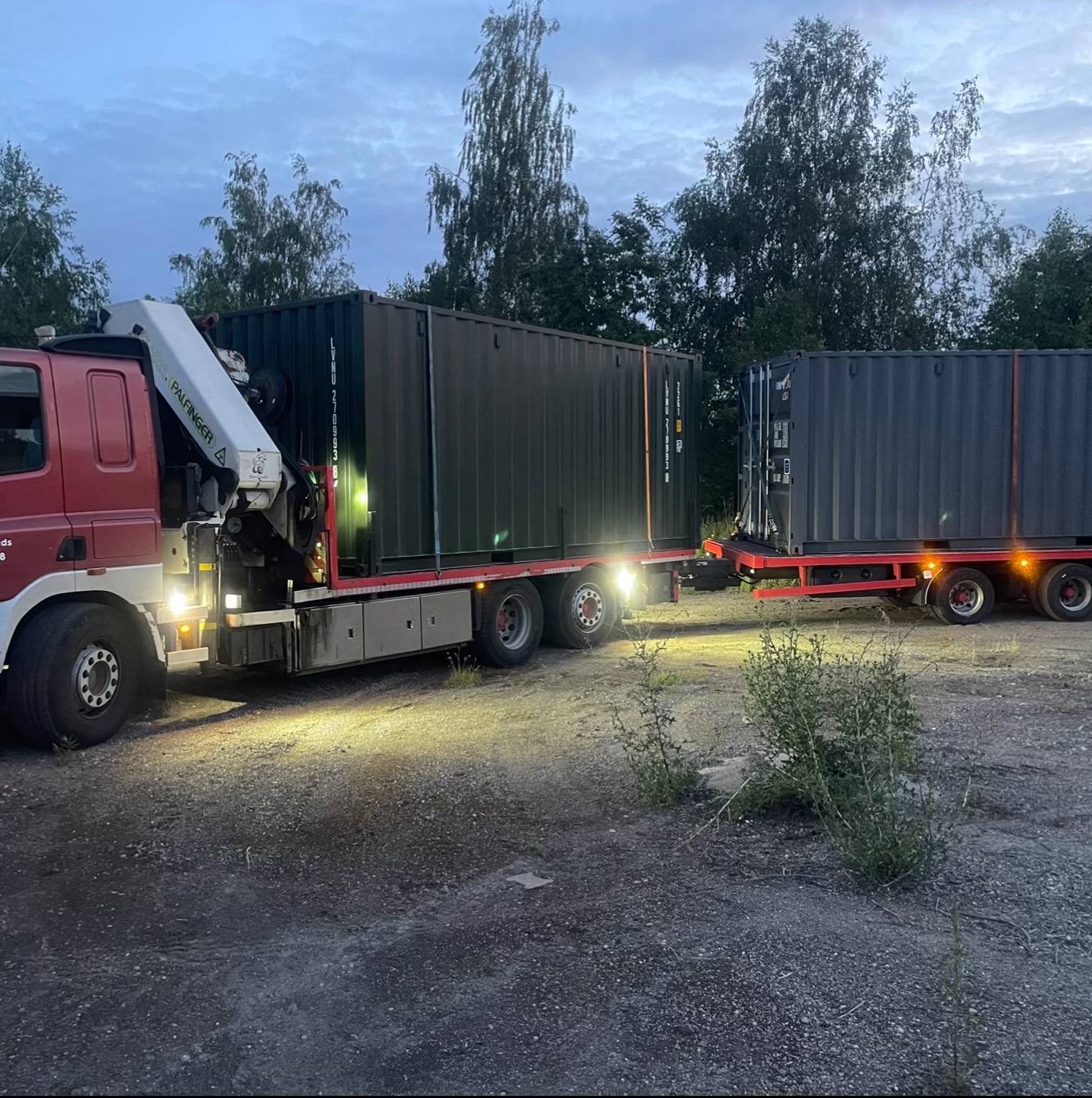 Czerwony dźwig samochodowy Palfinger transportuje dwa kontenery na naczepie w pochmurny dzień. Widoczne drzewa w tle.