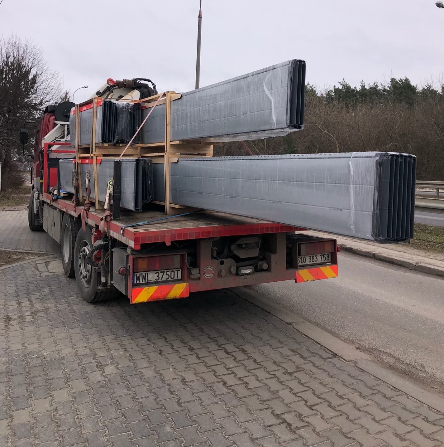 Czerwony samochód ciężarowy z ładunkiem stalowych elementów konstrukcyjnych, zabezpieczonych folią, w Zielonce. Transport specjalistyczny.