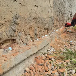 Iniekcja krystaliczna w zawilgoconej ścianie budynku. Widoczne nawiercone otwory wypełnione preparatem, kamienny fundament i dłoń w czerwonej rękawicy.