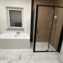 Nowoczesna łazienka z wanną i kabiną prysznicową typu walk-in. Białe marmurowe płytki podłogowe, ściana z drewnopodobnych paneli za kabiną. Minimalistyczny design.