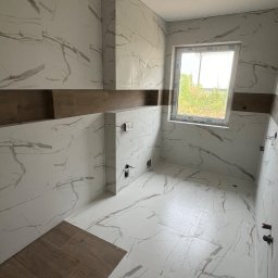 Pro House - Profesjonalne wykończenia wnetrz - Wnętrze łazienki w trakcie wykańczania, z białymi płytkami imitującymi marmur, brązowe akcenty dekoracyjne, okno z widokiem na zewnątrz, widoczne elementy instalacji.