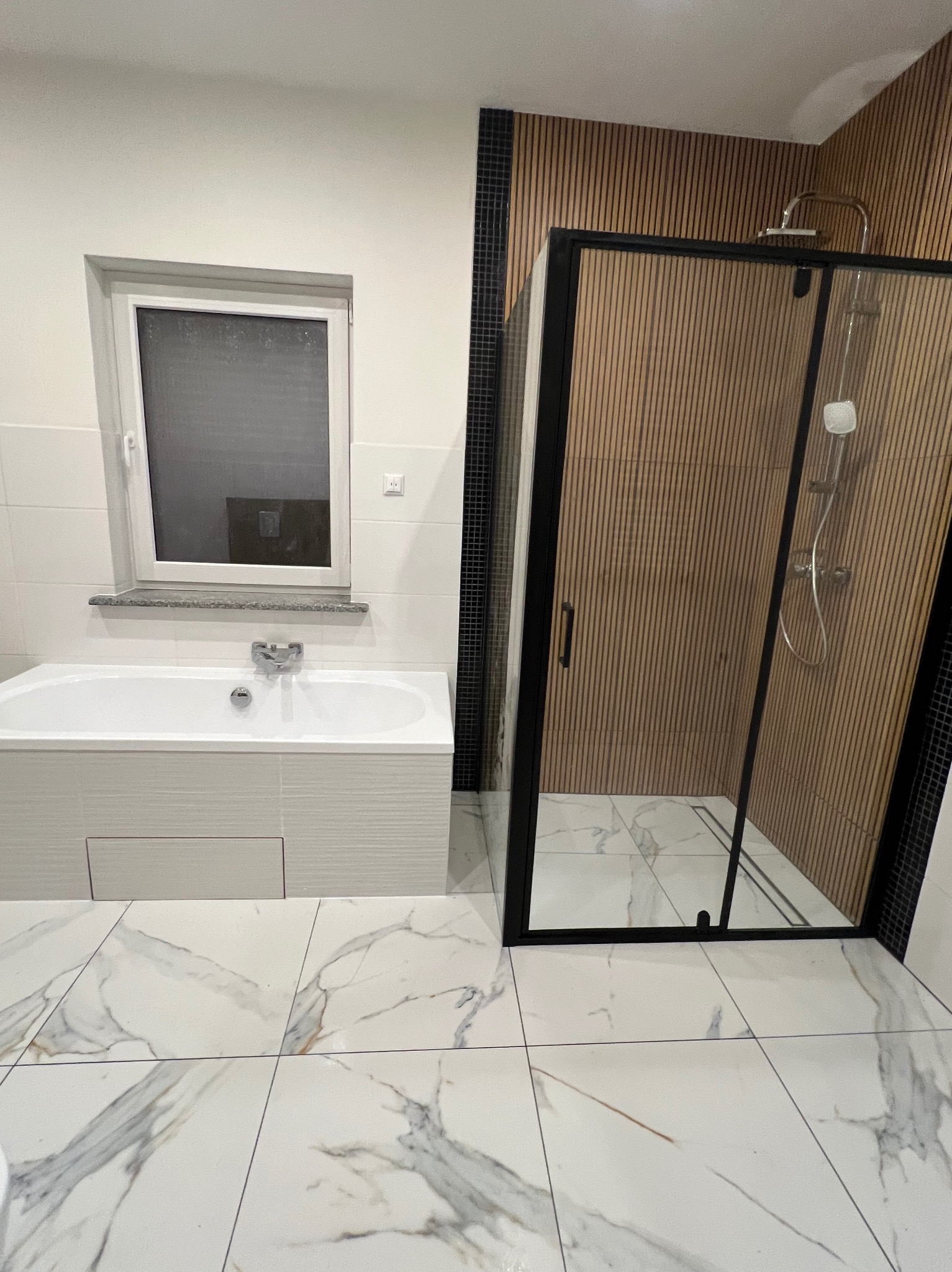 Nowoczesna łazienka z wanną i kabiną prysznicową typu walk-in. Białe marmurowe płytki podłogowe, ściana z drewnopodobnych paneli za kabiną. Minimalistyczny design.