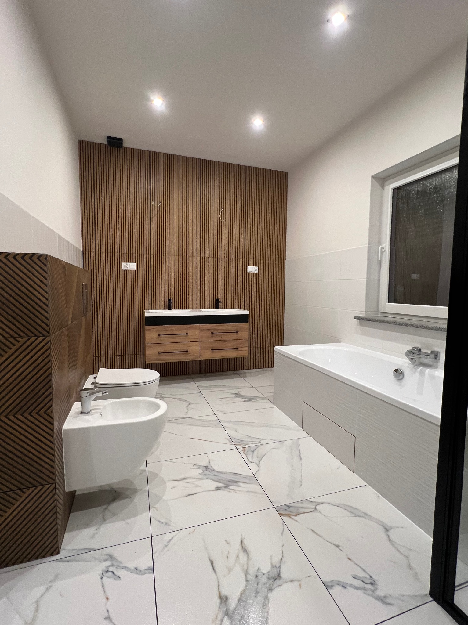 Nowoczesna łazienka z drewnianymi akcentami: panel ścienny, szafka pod umywalkę, płytki imitujące marmur. Bidet i toaleta po lewej stronie, wanna po prawej.
