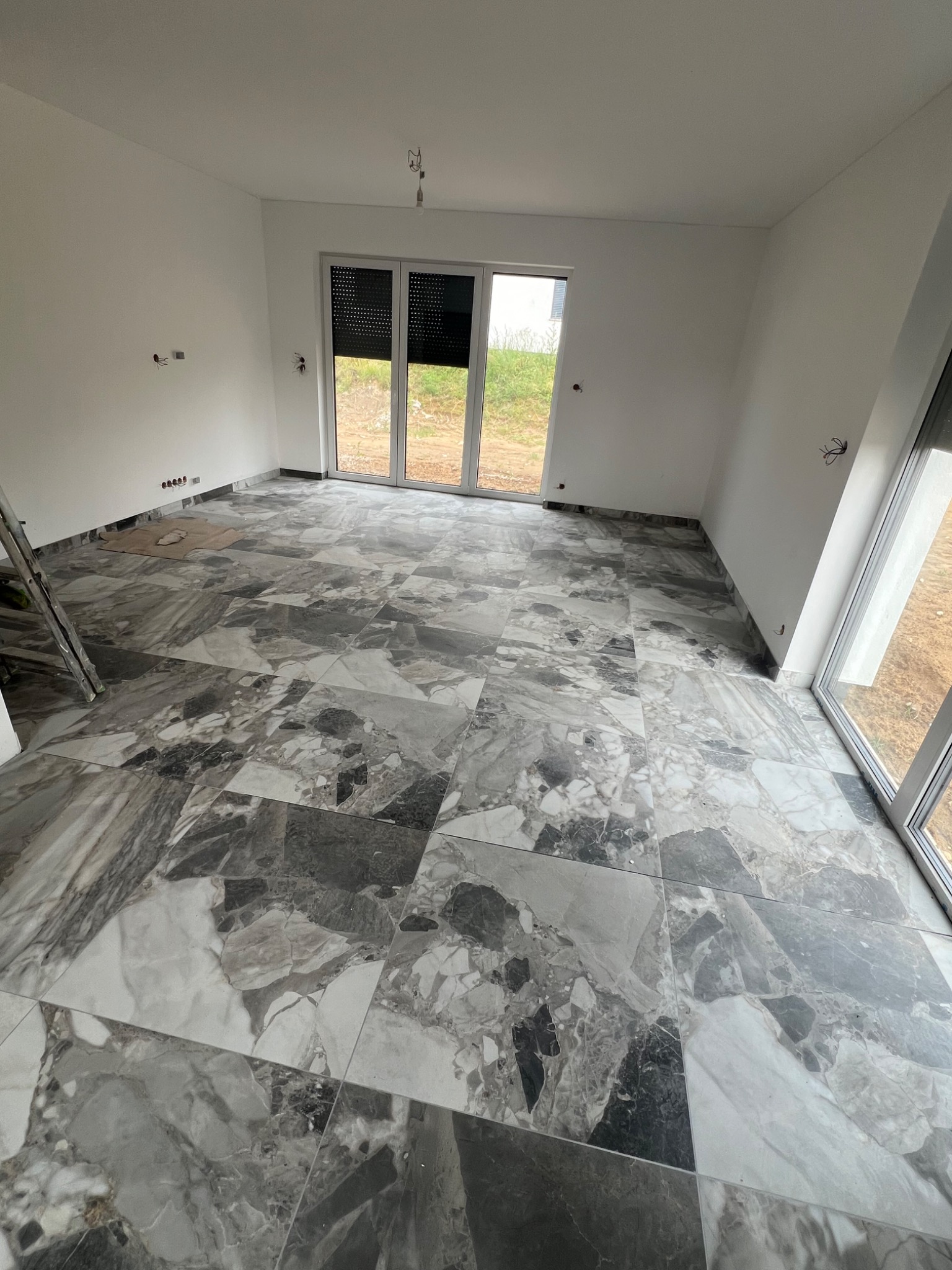 Ułożona podłoga z płytek imitujących marmur w jasnym pokoju z dużymi oknami balkonowymi. Widoczne fragmenty drabiny.