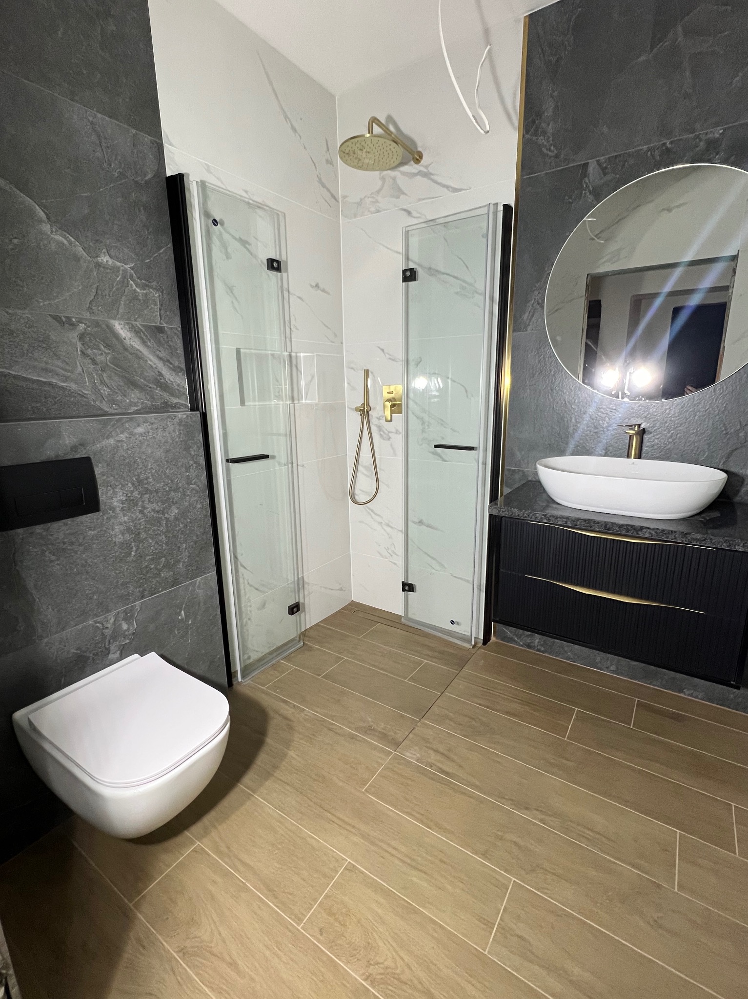 Nowoczesna łazienka z kabiną prysznicową typu walk-in, umywalką nablatową na czarnej szafce z złotymi uchwytami i toaletą podwieszaną. Dominuje szaro-biała kolorystyka.