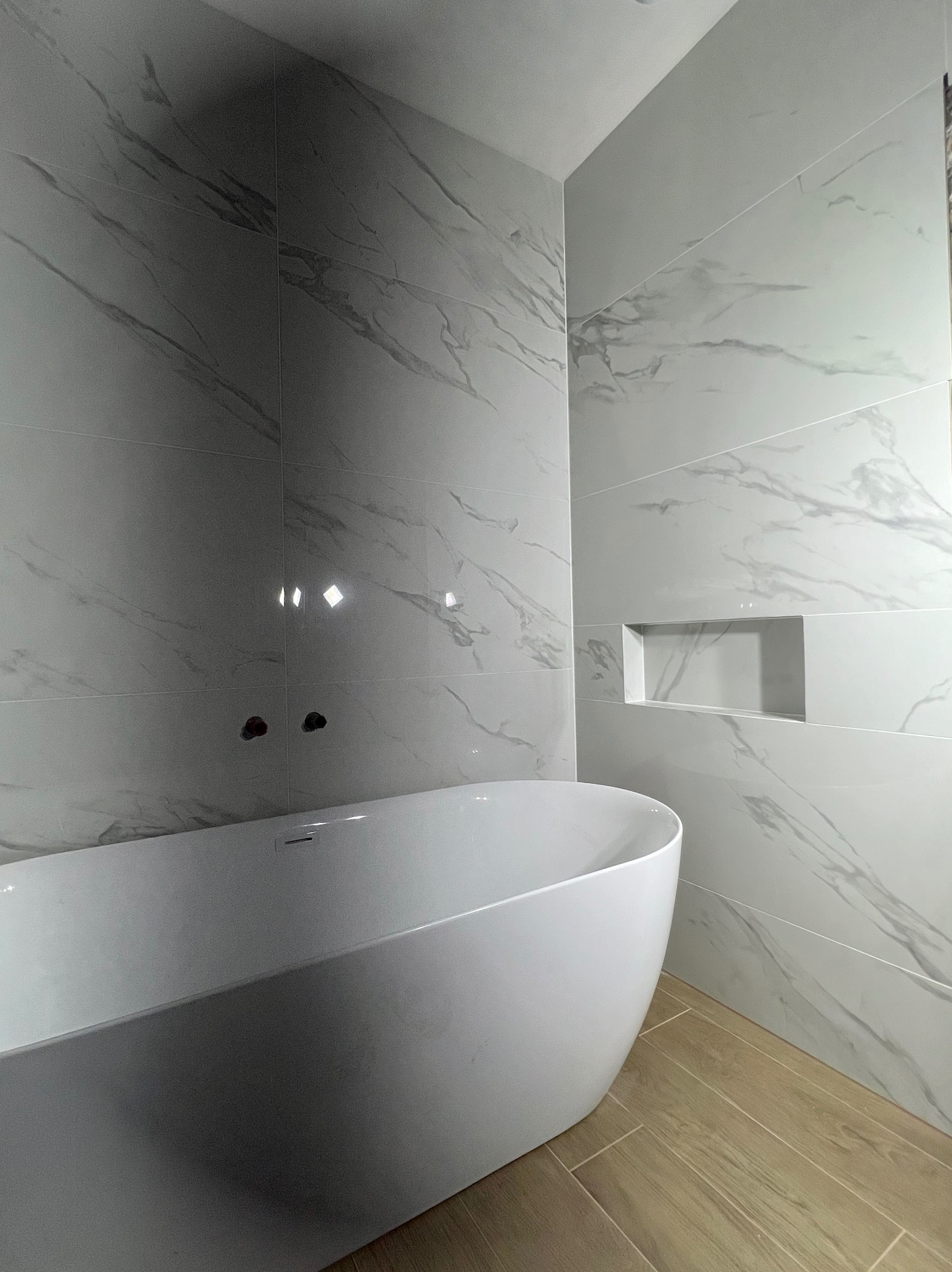 Nowoczesna łazienka z białą, wolnostojącą wanną i marmurowymi płytkami, akcentowana wnęką w ścianie. Podłoga wyłożona panelami imitującymi drewno.