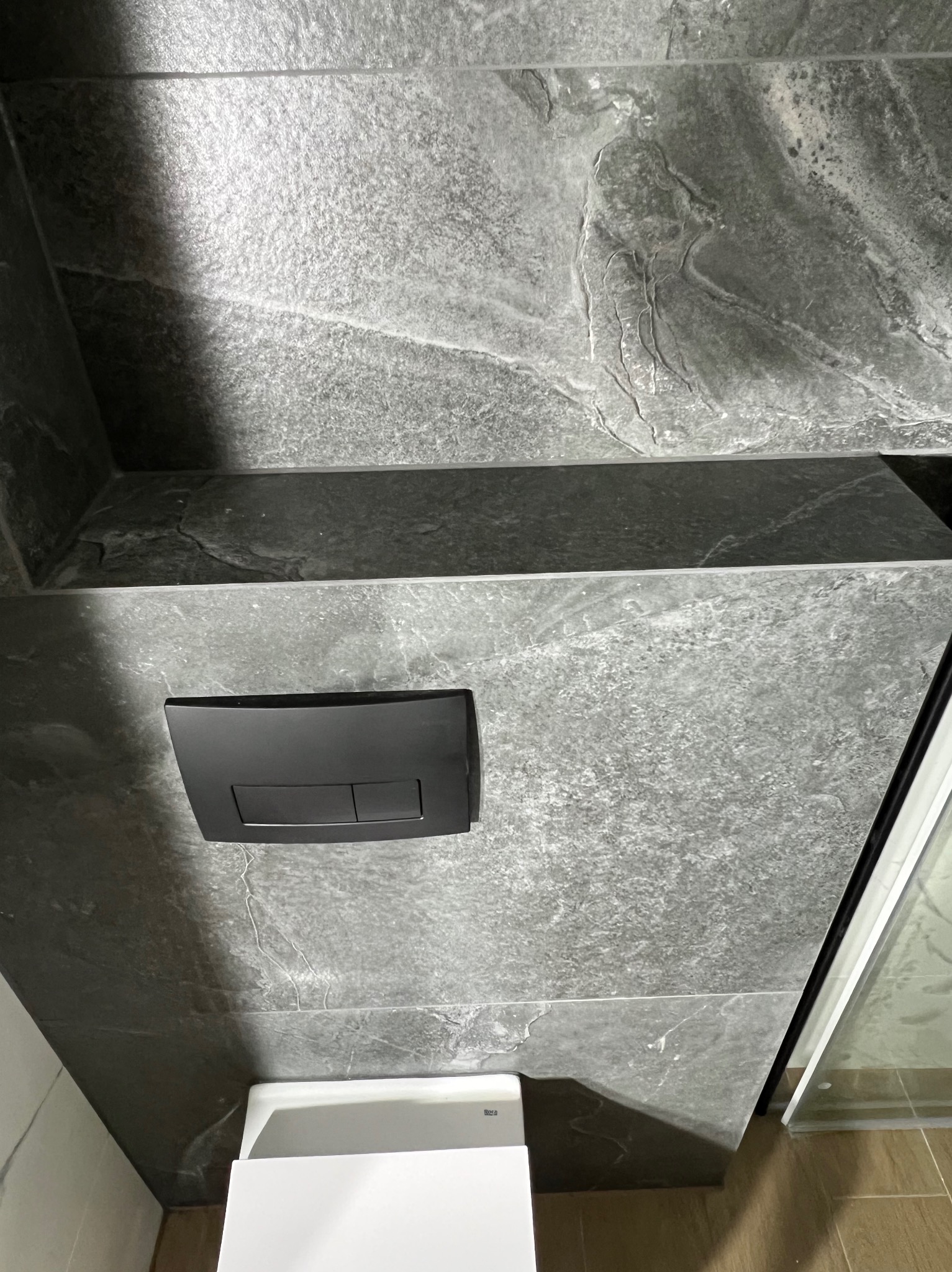 Wykończona łazienka z szarą, marmurkową płytką, czarnym przyciskiem spłuczki i częściowo widoczną białą toaletą. Nowoczesne i eleganckie wykończenie.