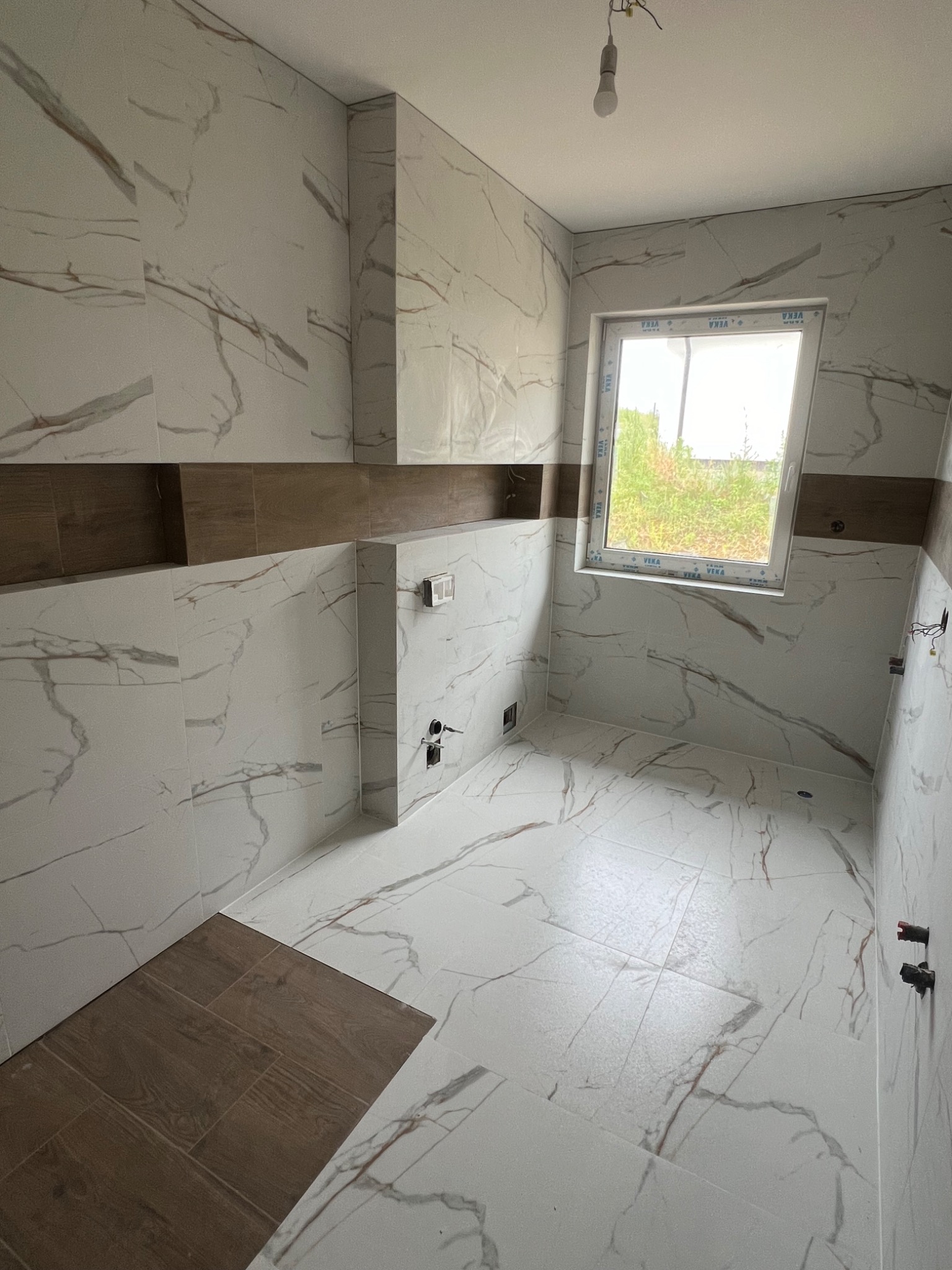Wnętrze łazienki w trakcie wykańczania, z białymi płytkami imitującymi marmur, brązowe akcenty dekoracyjne, okno z widokiem na zewnątrz, widoczne elementy instalacji.