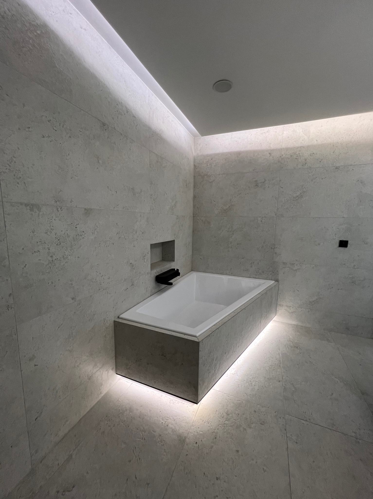 Minimalistyczna łazienka z wanną obudowaną szarą płytą, podświetloną od dołu. Ściany i podłoga wyłożone jasnoszarymi płytkami imitującymi kamień. Oświetlenie LED w suficie.