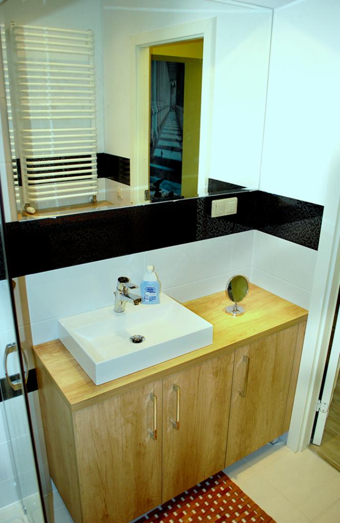 Nowoczesna łazienka z kwadratową umywalką nablatową, szafką z drewnopodobnym wykończeniem i lustrem z widocznym grzejnikiem drabinkowym w odbiciu.