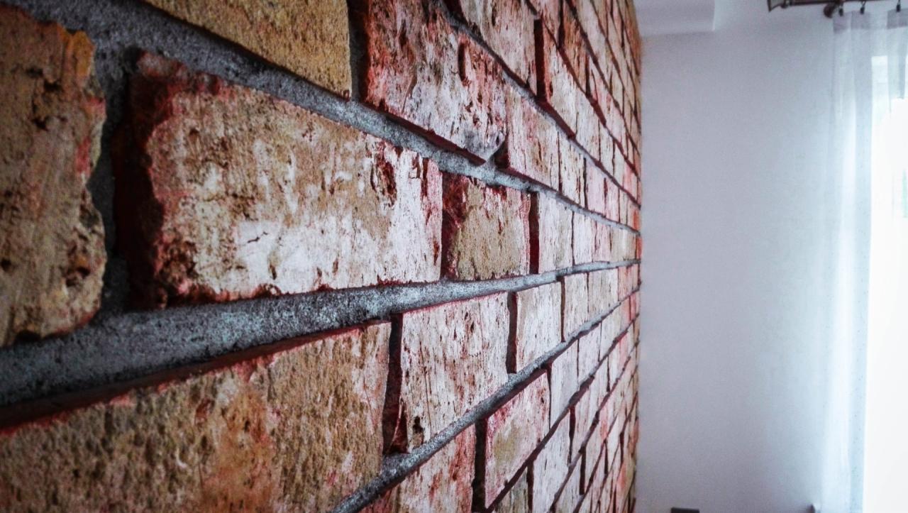 Dekoracyjna ściana z cegieł w odcieniach czerwieni i brązu z szarą fugą, widok z boku, z fragmentem białej ściany i zasłony po prawej stronie.