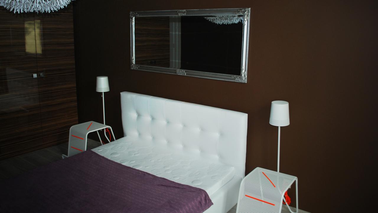 Nowoczesna sypialnia z białym łóżkiem z pikowanym zagłówkiem, lustrem w ozdobnej ramie na brązowej ścianie, minimalistycznymi lampkami nocnymi i szafkami z perforowanej blachy.