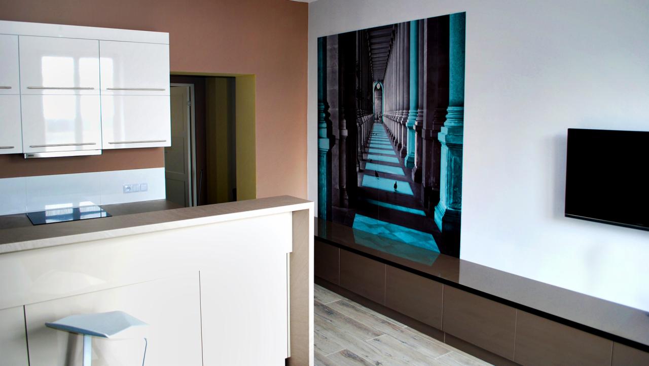 Nowoczesne wnętrze z aneksem kuchennym, białe szafki kuchenne, wyspa z hokerem, fototapeta z motywem kolumnady i telewizor na ścianie.