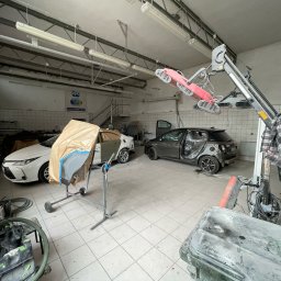 Lakard Sp z o.o. - Warsztat: biały samochód z osłoniętą karoserią, szary pojazd w trakcie naprawy, lampa grzewcza i narzędzia blacharskie w jasnym pomieszczeniu z białą glazurą.