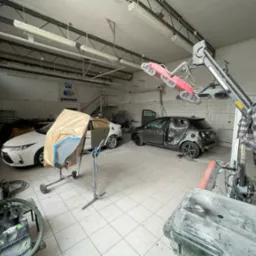 Warsztat: biały samochód z osłoniętą karoserią, szary pojazd w trakcie naprawy, lampa grzewcza i narzędzia blacharskie w jasnym pomieszczeniu z białą glazurą.