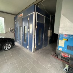 Lakard Sp z o.o. - Samochód z elementami w szarym podkładzie lakierniczym stoi przed kabiną lakierniczą Saima i kompresorem Airpress w warsztacie.