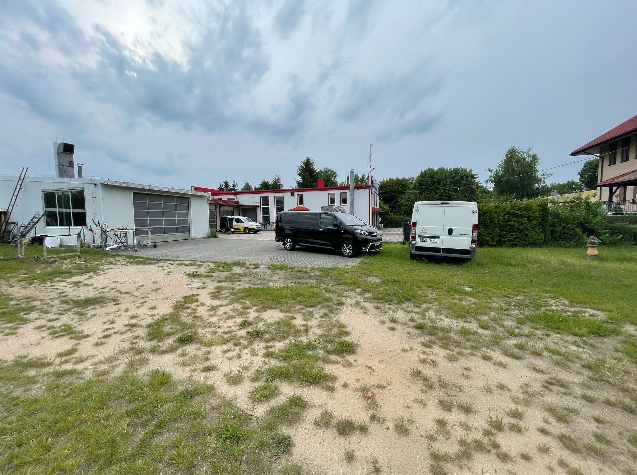 Warsztat samochodowy z zaparkowanymi pojazdami dostawczymi na placu. Widoczny budynek z bramą garażową i częściowo zarośnięty teren przed warsztatem.