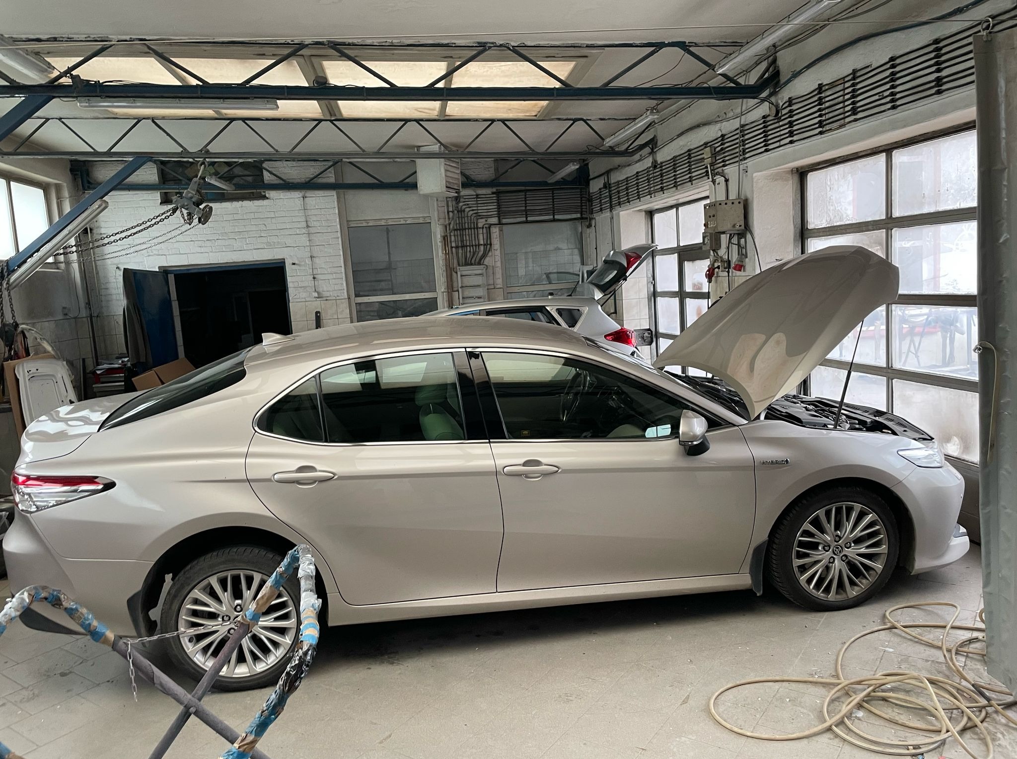 Beżowa Toyota Camry z otwartą maską w warsztacie; widoczny sprzęt blacharski i okna. Drugi samochód w tle, sufit z metalową konstrukcją.