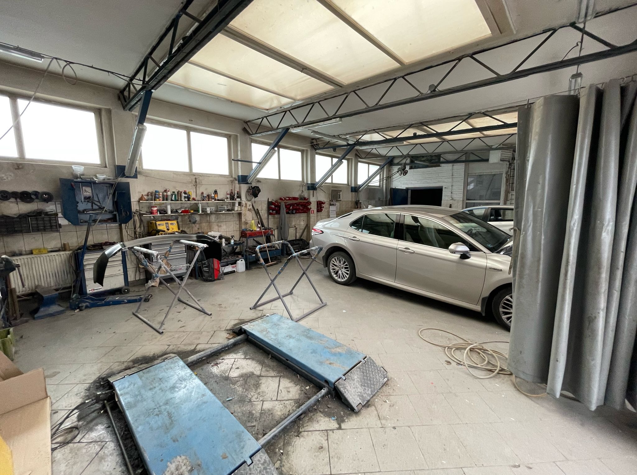 Wnętrze warsztatu: srebrny samochód na podnośniku, przygotowane narzędzia i elementy karoserii na stojakach, widoczne oświetlenie i konstrukcja dachu.